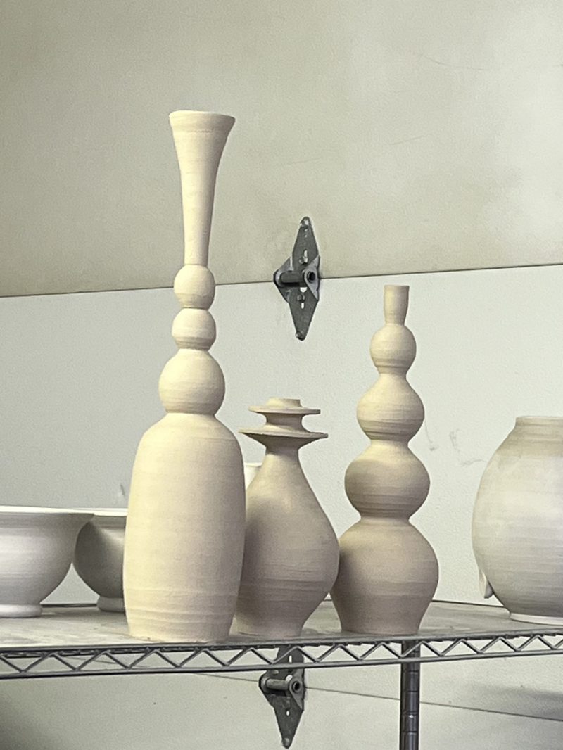 A tall flared bottle, a lidded bottle and a stacked-gourd form drying on a studio shelf.